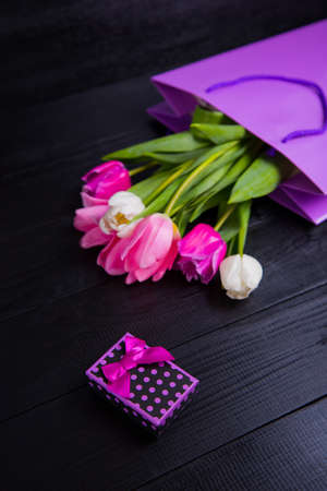Beautiful bouquet of tender pink and white tulips in purple shopping bag and gift box on black wooden background. Spring romantic composition.の写真素材