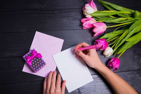 Female hands writing on clear paper, envelope, gift box and beautiful bouquet of tender pink and white tulips on black background. Spring romantic composition.の写真素材
