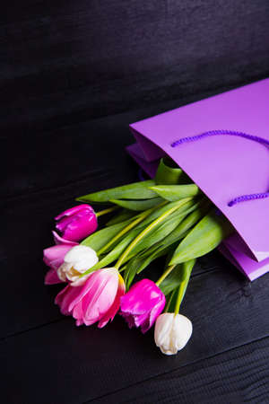 Beautiful bouquet of tender pink and white tulips in purple shopping bag on black wooden background. Spring romantic composition.の写真素材