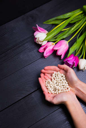 Beautiful bouquet of tender pink and white tulips with female hands holding pearl necklace in shape of heart on black wooden background. Spring romantic composition.の写真素材