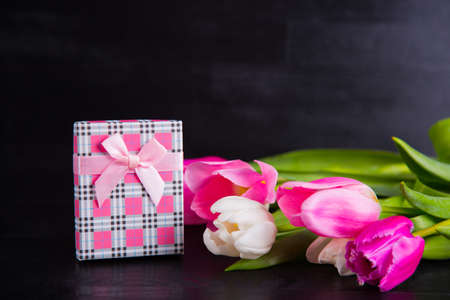 Beautiful bouquet of tender pink and white tulips and gift box on black wooden background. Spring romantic composition.の写真素材