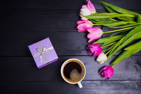 Cup of coffee, purple gift box and beautiful bouquet of tender pink and white tulips on black wooden background. Spring romantic composition.の写真素材