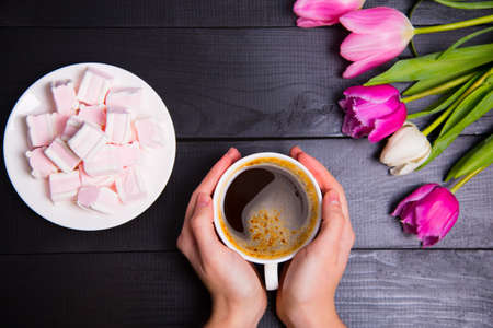 Beautiful bouquet of tender pink and white tulips with female hands holding cup of coffee and saucer with marshmallows on black wooden background. Spring romantic composition.の写真素材