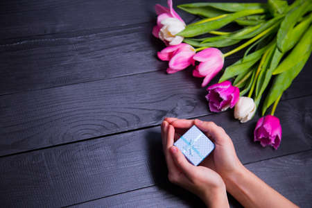 Beautiful bouquet of tender pink and white tulips with female hands holding gift box on black wooden background. Spring romantic composition.の写真素材