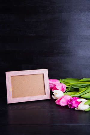 Beautiful bouquet of tender pink and white tulips and empty photo frame on black wooden background. Spring romantic composition.の写真素材