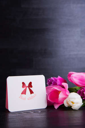 Beautiful bouquet of tender pink and white tulips with greeting card with inscription "Best wishes" on black wooden background. Spring romantic composition.の写真素材