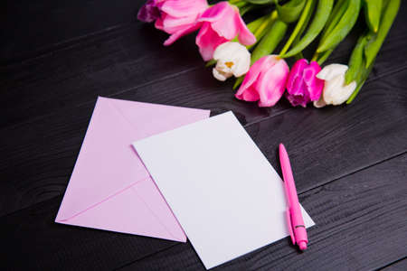 Beautiful bouquet of tender pink and white tulips, envelope, pen and clear paper sheet on black wooden background. Spring romantic composition.の写真素材