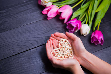 Beautiful bouquet of tender pink and white tulips with female hands holding wicker white heart on black wooden background. Spring romantic composition.の写真素材