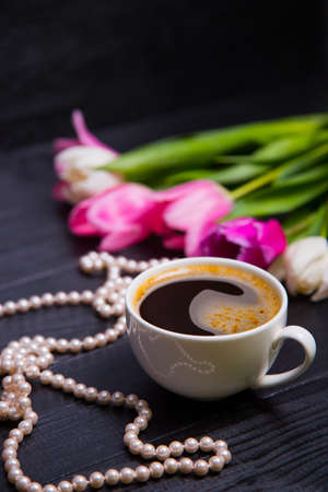 Cup of coffee, pearl necklace and beautiful bouquet of tender pink and white tulips on black wooden background. Spring romantic composition.の写真素材