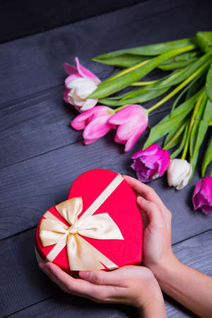 Beautiful bouquet of tender pink and white tulips with female hands holding gift box in shape of heart on black wooden background. Spring romantic composition.の写真素材