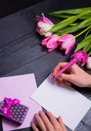 Female hands writing on clear paper, envelope, gift box and beautiful bouquet of tender pink and white tulips on black background. Spring romantic composition.の写真素材
