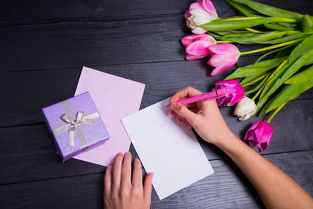 Female hands writing on clear paper, envelope, gift box and beautiful bouquet of tender pink and white tulips on black background. Spring romantic composition.の写真素材