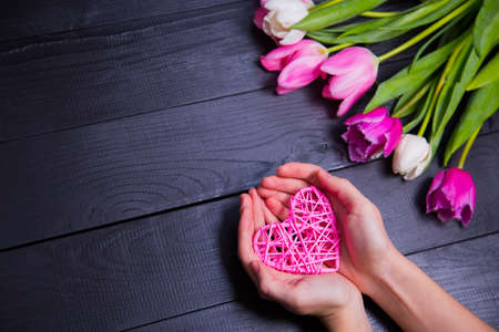 Beautiful bouquet of tender pink and white tulips with female hands holding wicker pink heart on black wooden background. Spring romantic composition.の写真素材