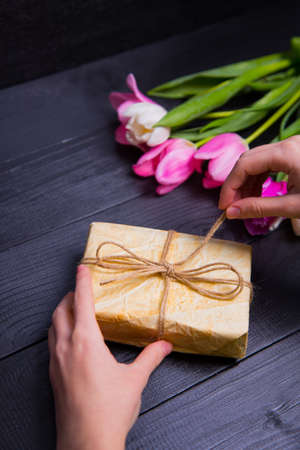 Beautiful bouquet of tender pink and white tulips with female hands packing and wrapping gift box on black wooden background. Spring romantic composition.の写真素材