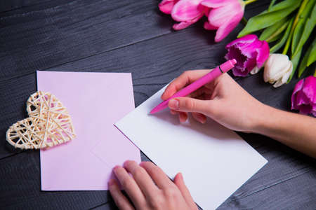 Female hands writing on clear paper, envelope, wicker heart and beautiful bouquet of tender pink and white tulips on black background. Spring romantic composition.の写真素材