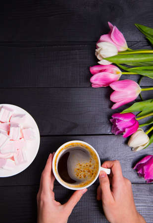 Beautiful bouquet of tender pink and white tulips with female hands holding cup of coffee and saucer with marshmallows on black woodの写真素材