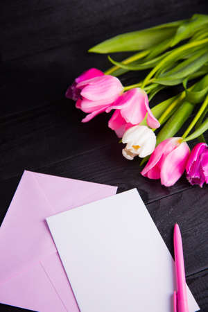 Beautiful bouquet of tender pink and white tulips, envelope, pen and clear paper sheet on black wooden background. Spring romantic composition.の写真素材