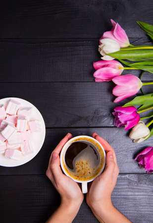Beautiful bouquet of tender pink and white tulips with female hands holding cup of coffee and saucer with marshmallows on black woodの写真素材