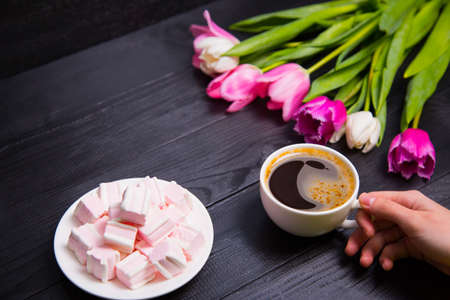 Beautiful bouquet of tender pink and white tulips with female hands holding cup of coffee and saucer with marshmallows on black woodの写真素材
