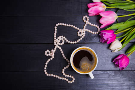 Cup of coffee, pearl necklace and beautiful bouquet of tender pink and white tulips on black woodの写真素材