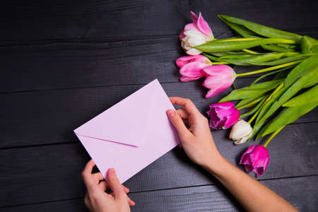 Beautiful bouquet of tender pink and white tulips with female hands holding pink envelope on black wooden background. Spring romantic composition.の写真素材