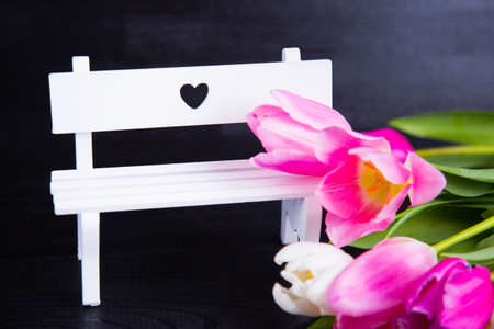 Beautiful bouquet of tender pink and white tulips and white bench with hole in form of heart on black woodの写真素材