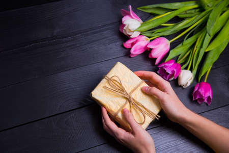 Beautiful bouquet of tender pink and white tulips with female hands holding gift box on black wooden background. Spring romantic composition.の写真素材