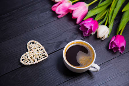 Cup of coffee, wicker white heart and beautiful bouquet of tender pink and white tulips on black wooden background. Spring romantic composition.の写真素材