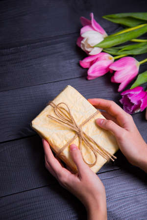 Beautiful bouquet of tender pink and white tulips with female hands holding gift box on black wooden background. Spring romantic composition.の写真素材