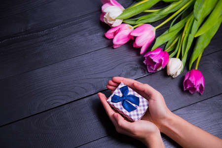 Beautiful bouquet of tender pink and white tulips with female hands holding gift box on black wooden background. Spring romantic composition.の写真素材