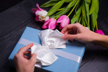 Beautiful bouquet of tender pink and white tulips with female hands packing and wrapping gift box on black wooden background. Spring romantic composition.の写真素材