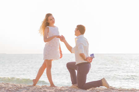Young romantic couple on seaside at golden sunrise. Man proposes to girlfriend on the beach.の写真素材