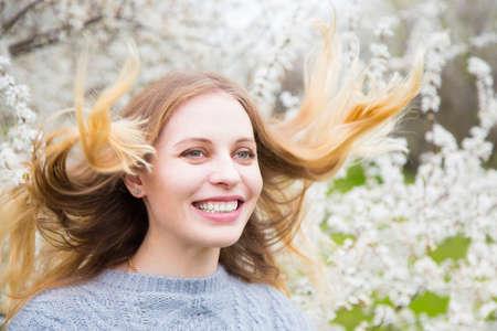 Portrait of beautiful blond young woman with fluttering hair standing in front of wonderful blooming trees in spring garden. Vintage colors.の写真素材