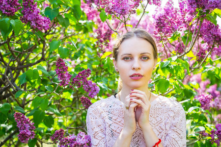 Close-up portrait of beautiful blond young woman standing between blooming lilac bushes in spring garden.の写真素材