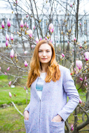 Close-up portrait of beautiful red-haired young woman standing near blooming magnolia branches in spring garden.の写真素材