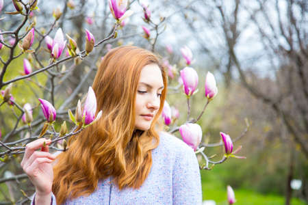 Close-up portrait of beautiful red-haired young woman standing near blooming magnolia branches in spring garden.の写真素材