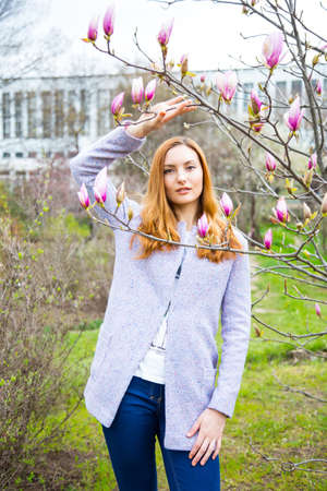 Close-up portrait of beautiful red-haired young woman standing near blooming magnolia branches in spring garden.の写真素材