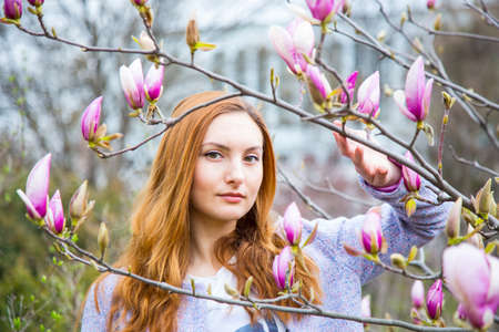 Close-up portrait of beautiful red-haired young woman standing near blooming magnolia branches in spring garden.の写真素材