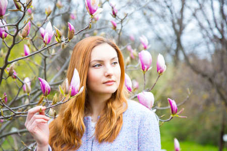 Close-up portrait of beautiful red-haired young woman standing near blooming magnolia branches in spring garden.の写真素材