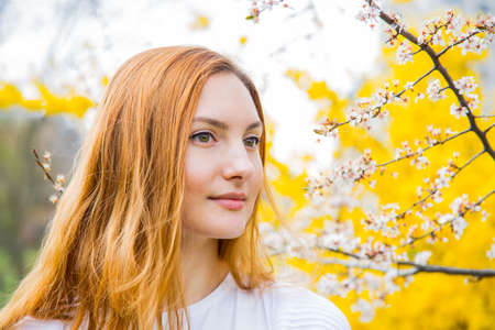 Close-up portrait of beautiful red-haired young woman standing near blooming cherry branches in spring garden.の写真素材