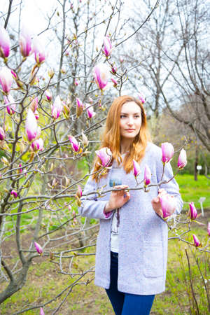 Close-up portrait of beautiful red-haired young woman standing near blooming magnolia branches in spring garden.の写真素材