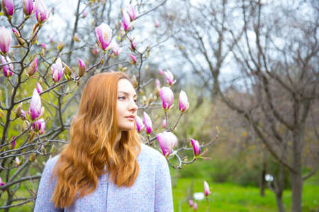 Close-up portrait of beautiful red-haired young woman standing near blooming magnolia branches in spring garden.の写真素材