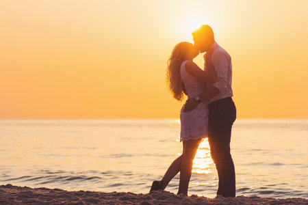 Beautiful young romantic couple kissing on seaside in rays of rising sun. Two happy lovers at golden sunrise. Warm colors.の写真素材