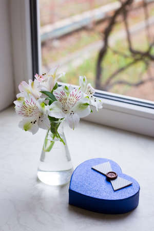 Beautiful tender bouquet of white Alstroemeria in glass vase with gift box in shape of heart near window. Spring composition. Springtime. Mother`s day card.の写真素材