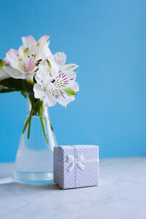 Beautiful tender bouquet of white Alstroemeria in glass vase with white gift box on blue background. Spring composition. Springtime. Mother`s day card.の写真素材