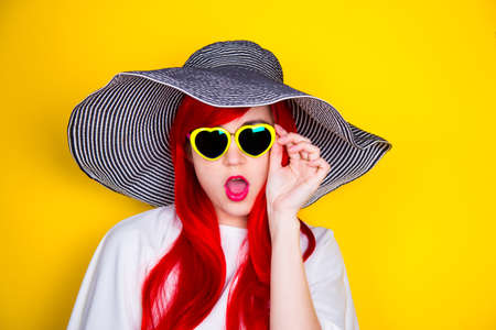 Attractive surprised red-haired young woman in sunglasses in form of heart and striped white and black hat on yellow summer background. Sexy stylish model enjoying sun.の写真素材