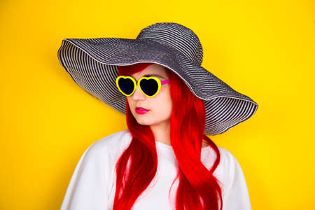 Attractive red-haired young woman in sunglasses in form of heart and striped white and black hat on yellow summer background. Sexy stylish model enjoying sun.の写真素材