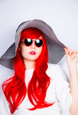 Attractive red-haired young woman in sunglasses in form of heart and striped white and black hat on white background. Sexy stylish model enjoying sun. Summertime.の写真素材