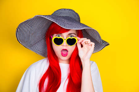 Attractive surprised red-haired young woman in sunglasses in form of heart and striped white and black hat on yellow summer background. Sexy stylish model enjoying sun.の写真素材