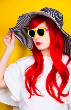 Attractive red-haired young woman in sunglasses in form of heart and striped white and black hat on yellow summer background. Sexy stylish model enjoying sun.の写真素材